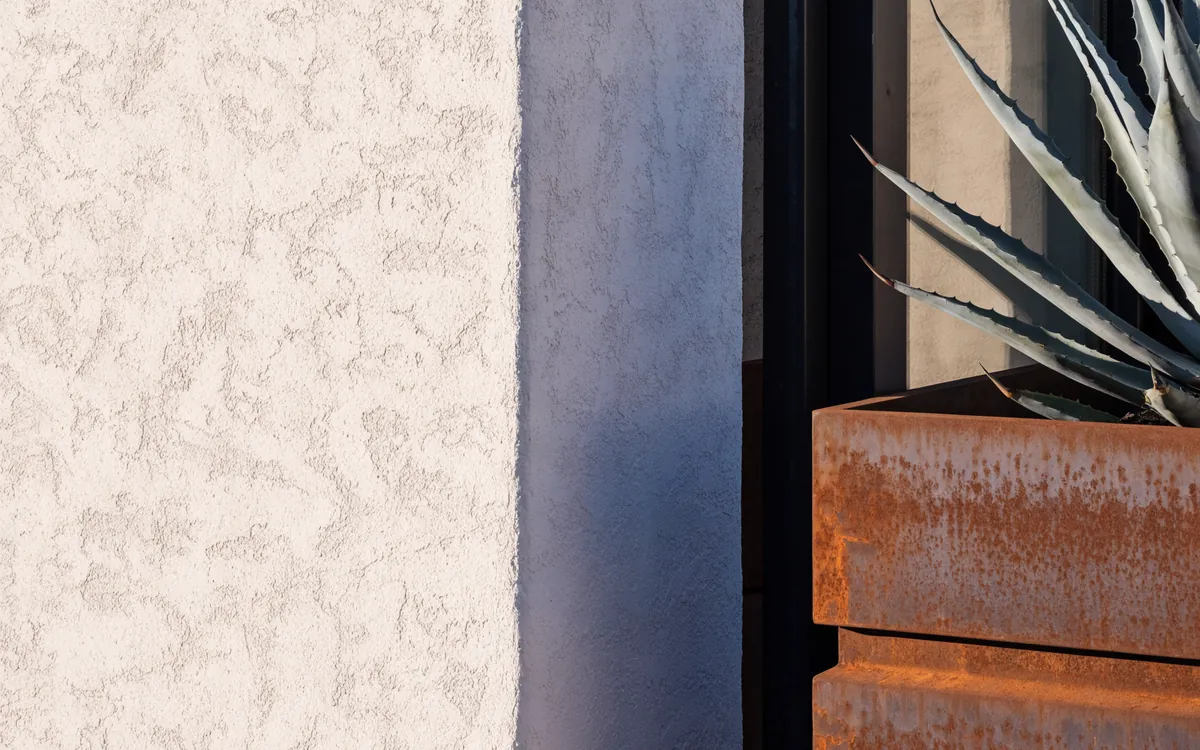 Close detail: hand-troweled white stucco, a blackened steel window frame and an agave in a weathered steel planter, in late sun.