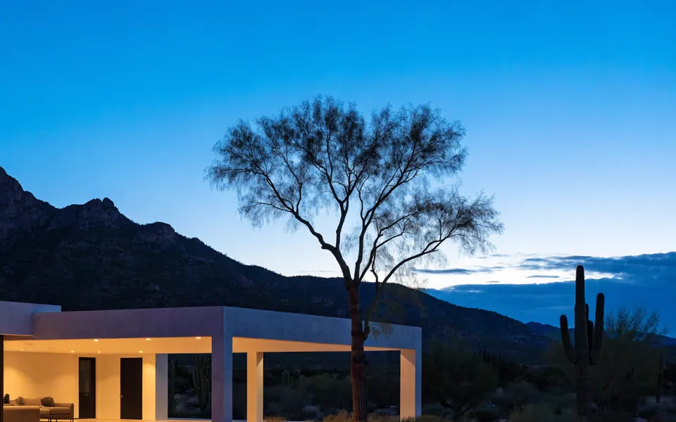 Blue hour over the site: a ridge of desert mountains, a lone mesquite tree and the edge of the porch roof.