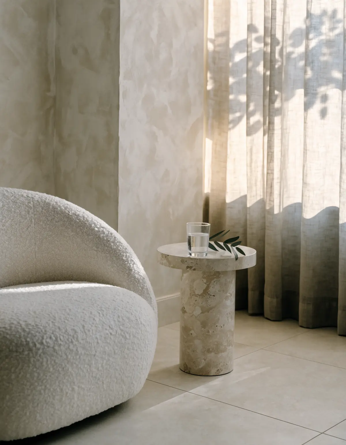 The lounge in late afternoon: a round boucle chair beside a travertine side table with a glass of water and an olive sprig, linen curtains lit by the sun.