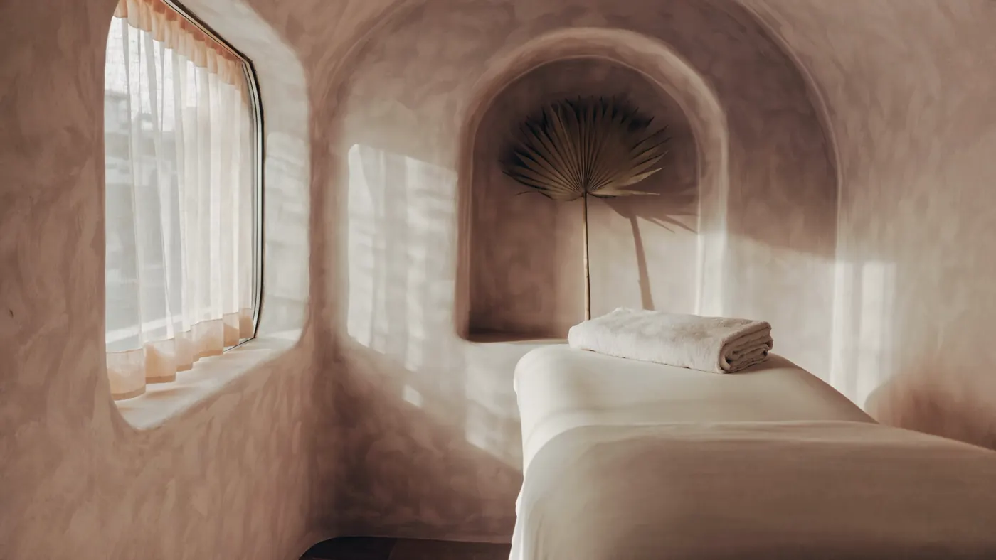 The treatment room: a curved plaster window with sheer curtains moving in the light, a linen-covered bed with a folded towel, and a dried palm frond in an arched niche.