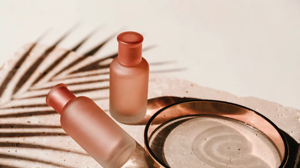 Two frosted rose glass bottles on travertine beside a brass dish, the shadow of a palm frond moving slowly across them.