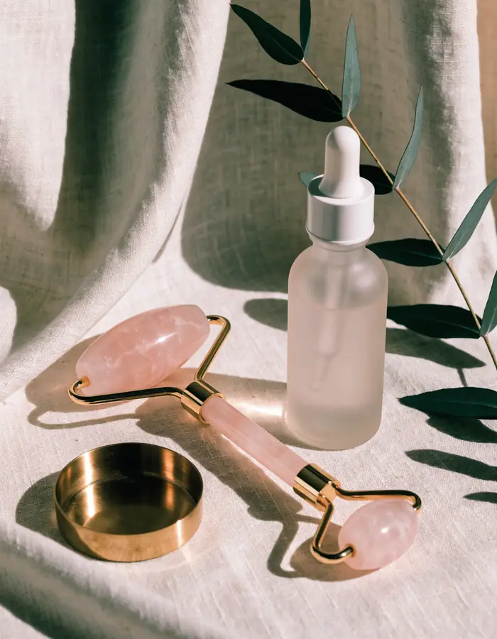 A rose quartz facial roller and a frosted glass dropper bottle on draped linen in the sun, a eucalyptus sprig behind them.