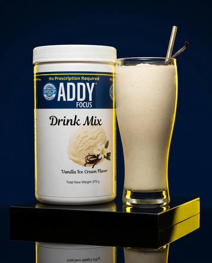 A tub of ADDY Focus drink mix beside a tall glass of vanilla shake on a black plinth.